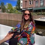 A woman wearing sunglasses and a colorful shirt is smiling while sitting in a white boat on a sunny day. She is enjoying a leisurely boat ride along an Amsterdam canal with quaint buildings and a bridge in the background, capturing the essence of a relaxed Dutch urban experience.