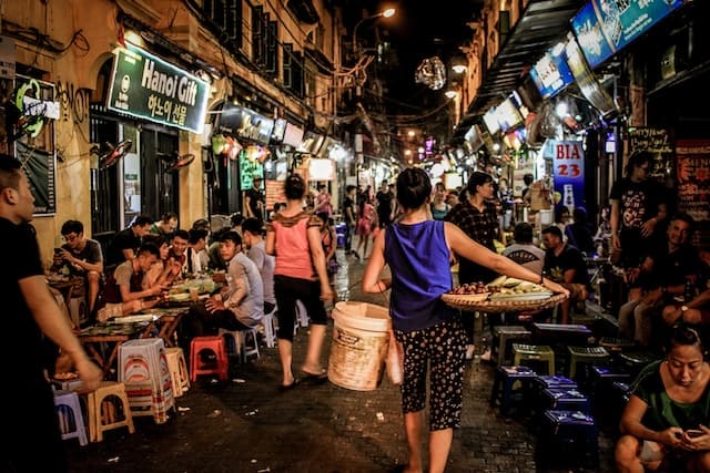 Vibrant street scene at night in Hanoi, Vietnam, bustling with locals dining at small tables along a narrow alley. A vendor in the foreground carries a large bamboo basket and tray, indicative of the lively street food culture, while neon signs and the glow of the market create a lively atmosphere.