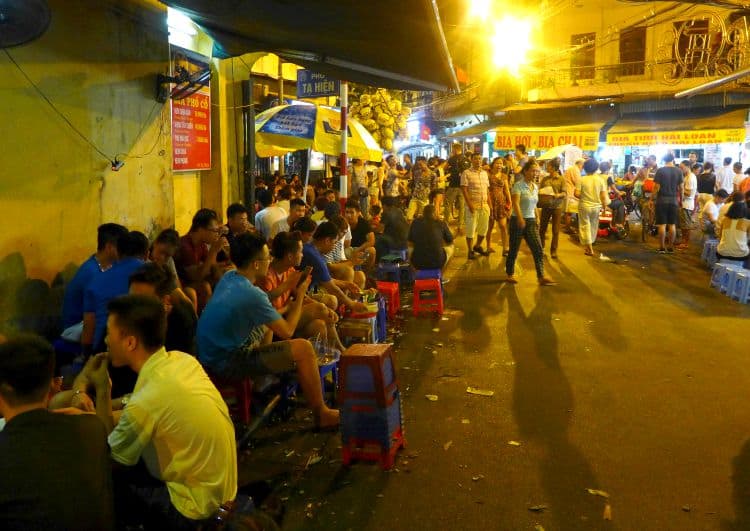 A lively street scene at night in Hanoi with rows of people seated on small plastic stools at makeshift outdoor eateries. The warm glow of streetlights illuminates the area, highlighting signs for 'Bia Hoi' and 'Bia Cafe,' as pedestrians and motorbikes navigate through the bustling urban nightlife.