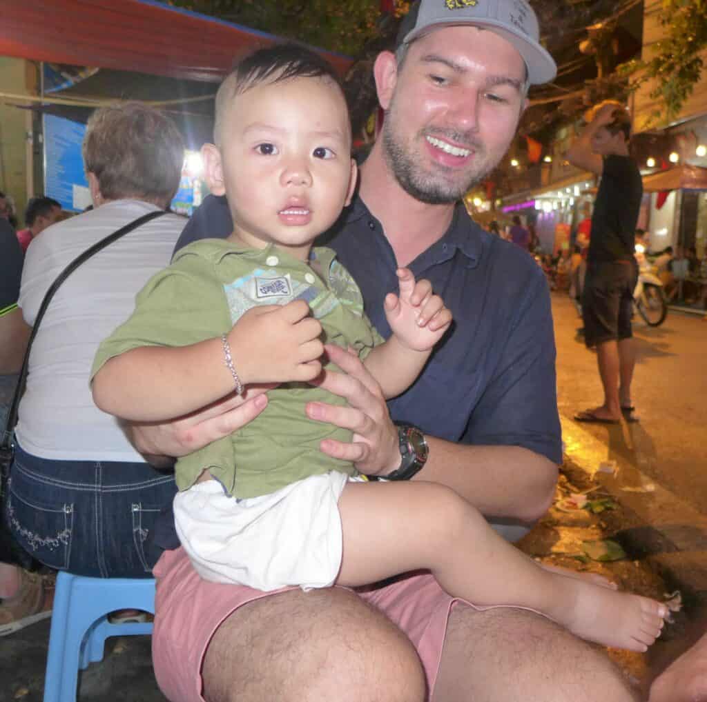 A smiling man wearing a baseball cap and dark shirt holds a young child with a surprised expression. The child is wearing a green shirt and has a bracelet on his wrist. They are seated, with the bustling street of a night market in Hanoi blurred in the background.