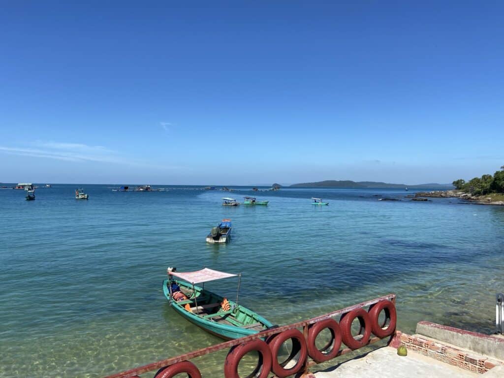 A peaceful coastal scene in Phu Quoc, Vietnam with multiple traditional boats floating on clear turquoise waters. A red railing with circular cut-outs lines the foreground, framing the view of the calm sea and a hilly island in the distance under a bright blue sky.