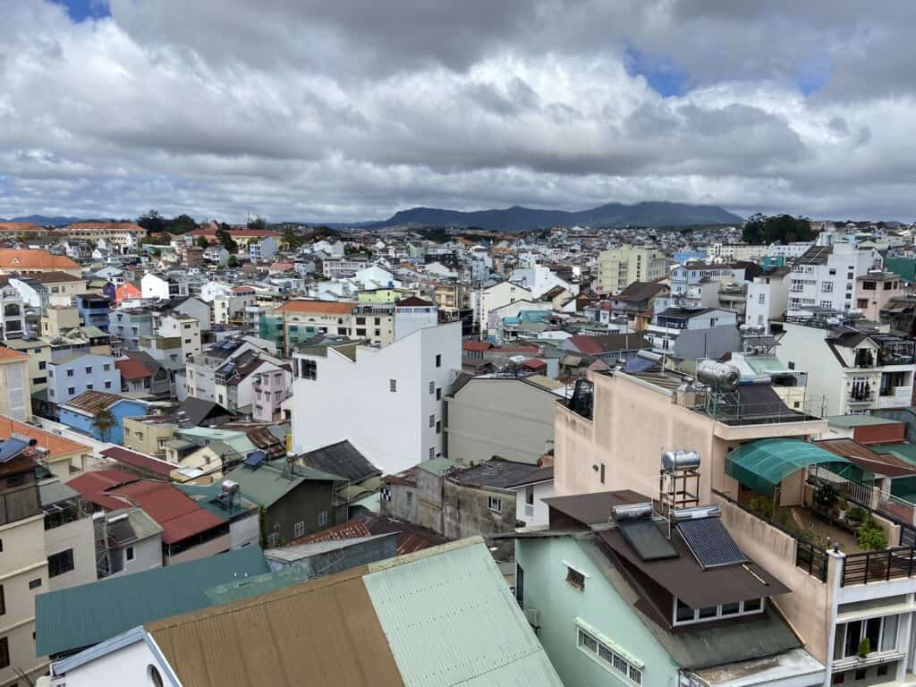 Overlooking a dense tapestry of residential and commercial buildings in Dalat, VIetnam, the photograph captures the urban landscape's varied architecture under a cloudy sky, with mountains faintly visible in the background.