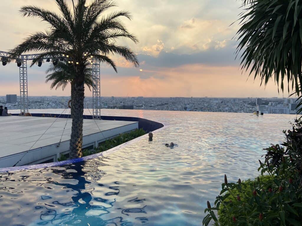 A rooftop infinity pool overlooks a bustling cityscape at sunset, with the sky painted in shades of orange and blue. Palm trees in the foreground add a tropical touch to this urban oasis, highlighting a serene escape amidst the city's expanse