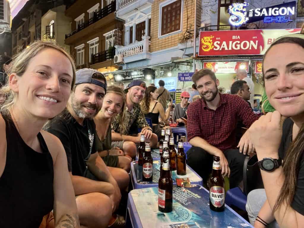 A group of six cheerful friends taking a selfie at an outdoor bar. They are seated at a blue table with several bottles of Bia Viet beer in front of them. The background features illuminated signs, one of which reads 'Saigon Grill,' contributing to the casual and friendly nightlife atmosphere of the setting.