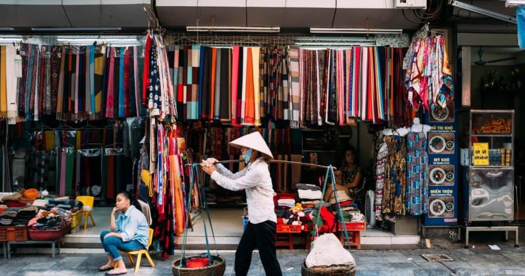 A Vietnamese textile shop displays a colorful array of fabrics and scarves while a person wearing a traditional conical hat walks by carrying a shoulder pole with baskets, exemplifying the vibrant street life of Vietnam.
