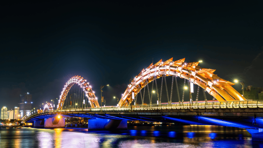 The Dragon Bridge in Da Nang, Vietnam, illuminated at night with glowing lights outlining its unique dragon-shaped design. The bridge arches majestically over the calm waters of the river, while the city's skyline, lit by streetlights and urban glow, stretches into the distance under the night sky