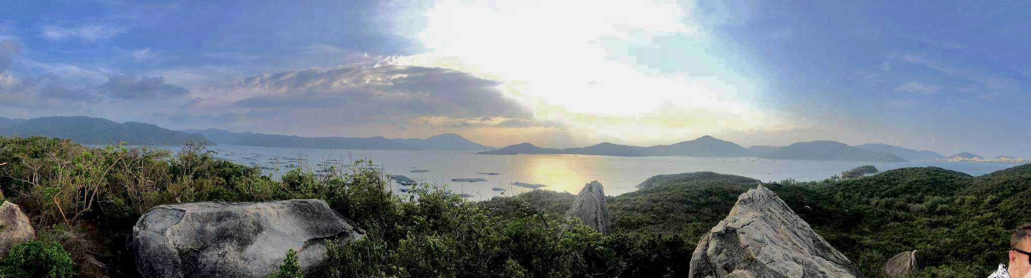 Panoramic view of a serene coastal landscape at sunset, showcasing a wide expanse of sea bordered by lush, green hills. The sky, partly cloudy, reflects the sun's golden hues onto the water's surface, with distant mountains and islands adding depth to the tranquil scene.