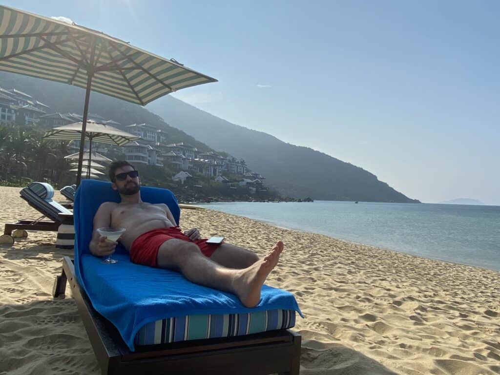 A relaxed man in sunglasses and red swim trunks lounges on a beach chair under a striped umbrella, holding a cocktail, with a serene beach and hillside villas in the background. The clear blue sky and calm sea create a perfect tropical setting for relaxation and leisure.