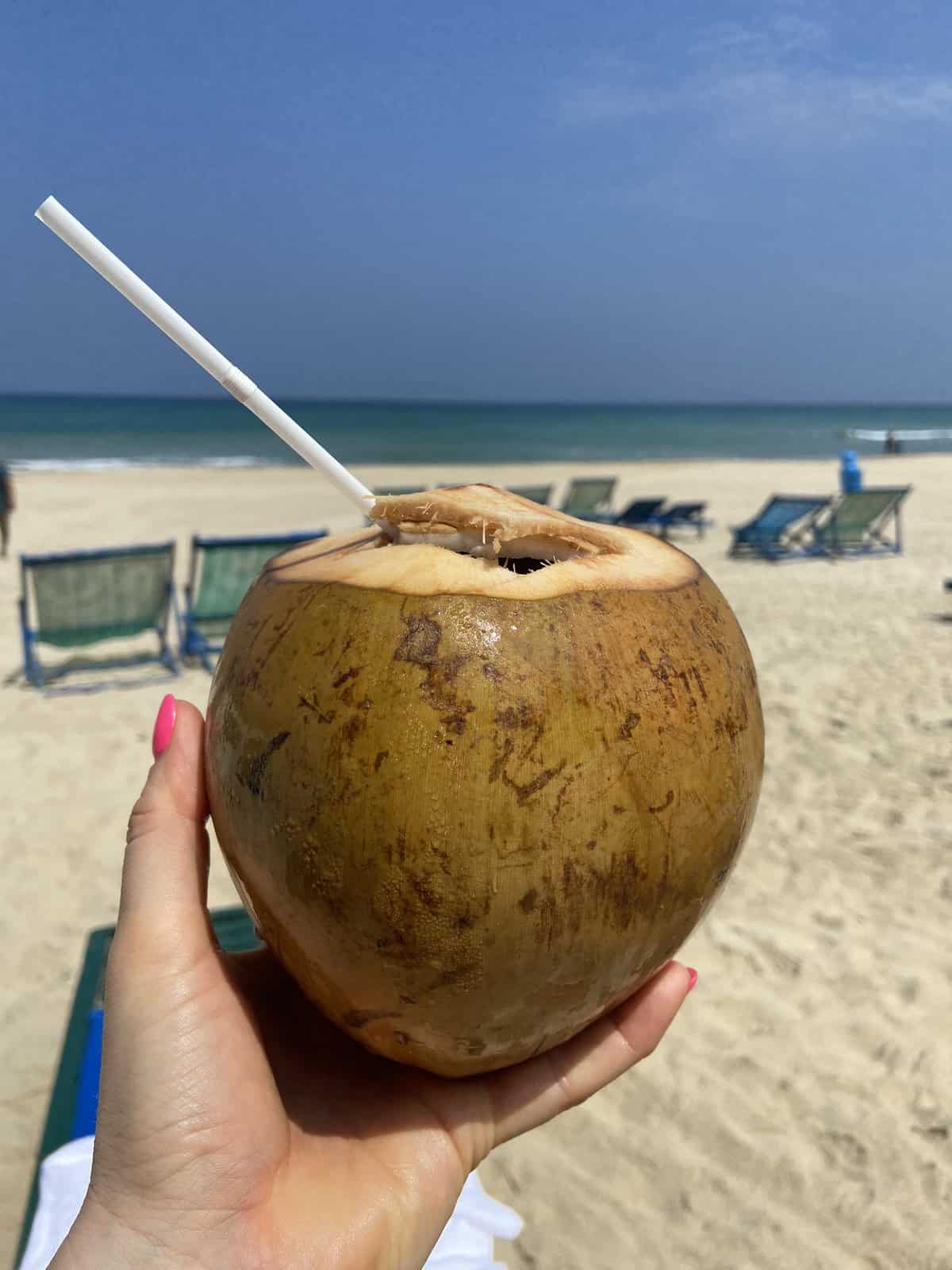 A hand holding a fresh coconut with a straw against a sunny beach backdrop, with beach chairs on the sand and the clear blue ocean in the distance