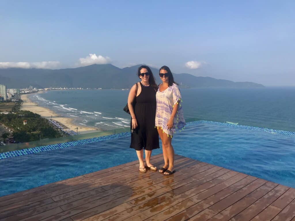 Two smiling women standing together on the edge of an infinity pool, with a panoramic view of a long sandy beach, ocean waves, and mountainous coastline in the background, under a clear blue sky.