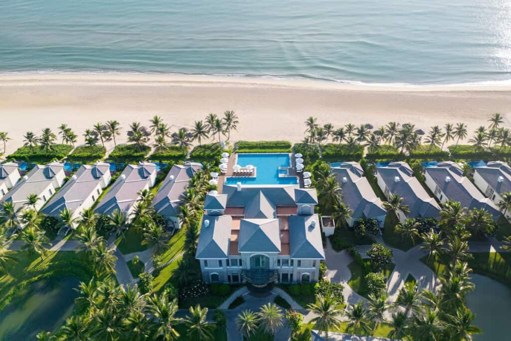Aerial view of a luxurious beachside resort with a central mansion flanked by rows of villas, a large swimming pool, and palm trees, leading up to a sandy beach and the ocean.