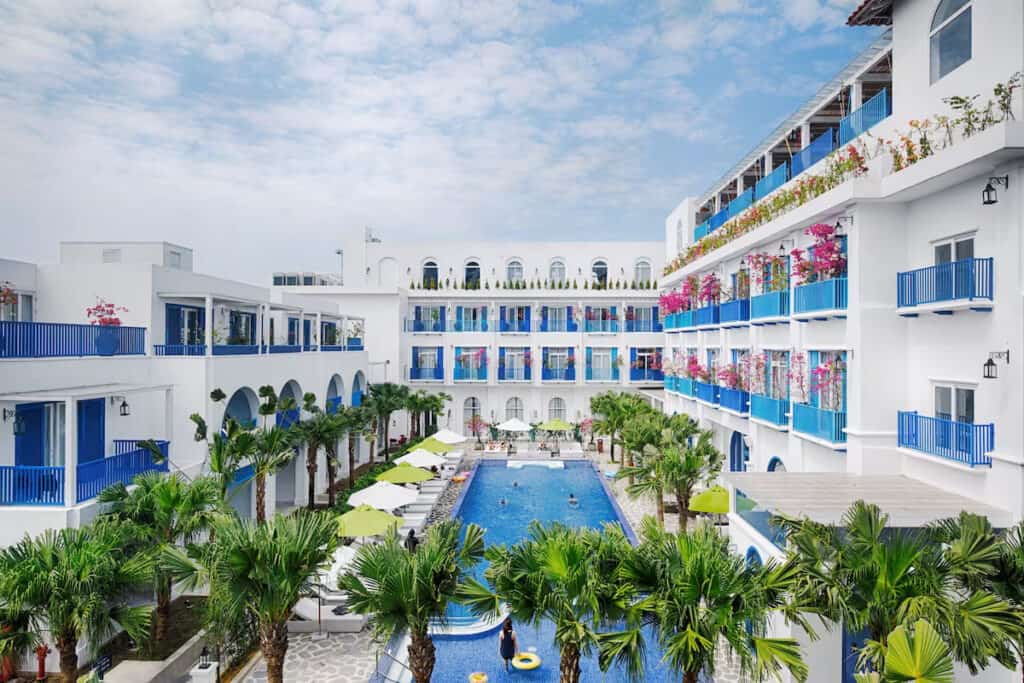Courtyard view of a Mediterranean-inspired resort with white buildings featuring blue railings and colorful bougainvillea, surrounding a central swimming pool with loungers and umbrellas.
