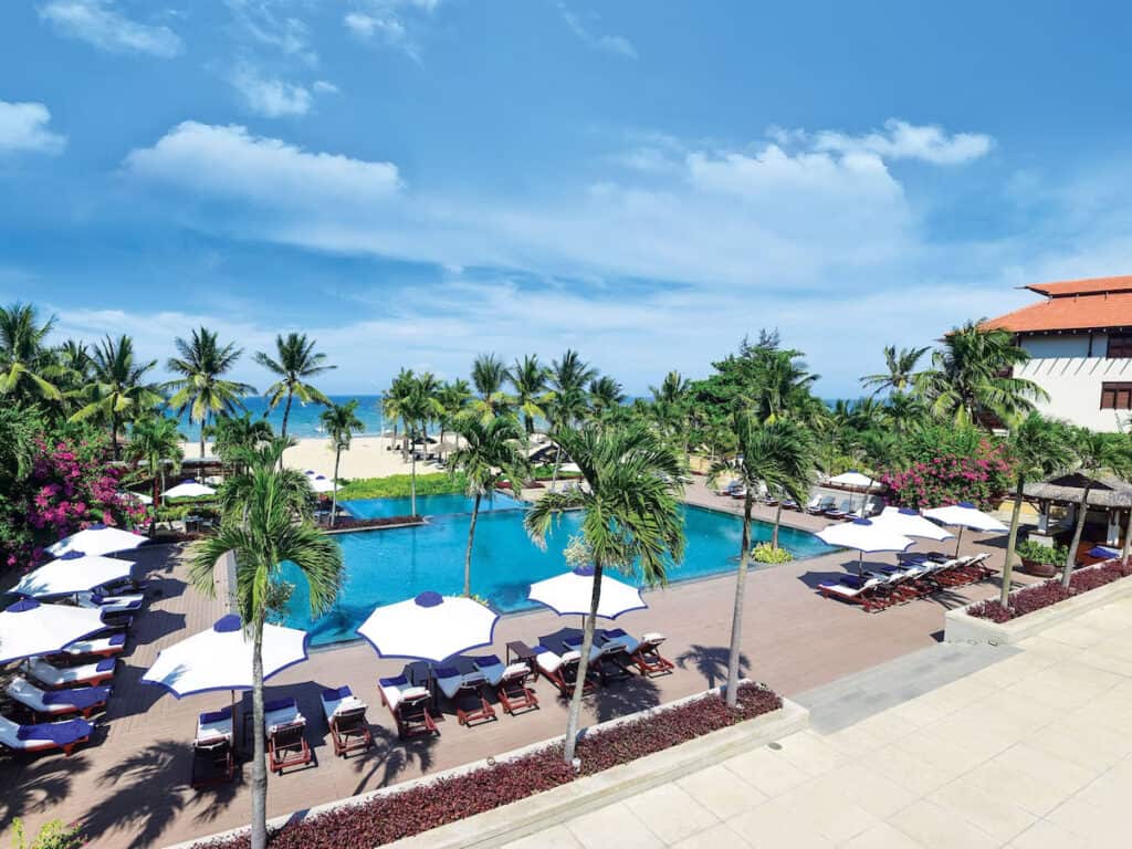 Resort poolside view with white sun umbrellas and loungers lined up on a wooden deck, surrounded by tropical palm trees, with a sandy beach and clear blue sky in the background