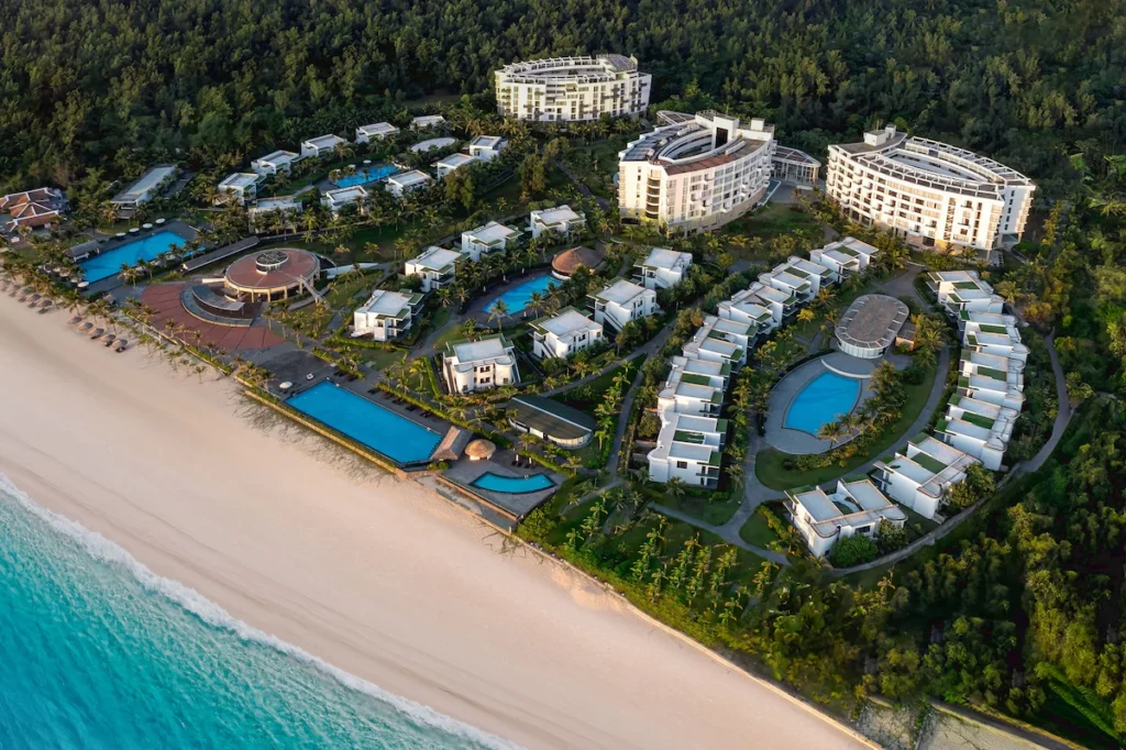 wilight descends over a luxurious beachfront resort complex, with modern white buildings surrounded by tropical foliage, multiple pools glistening near the sandy shore, and the ocean's vast expanse in the background