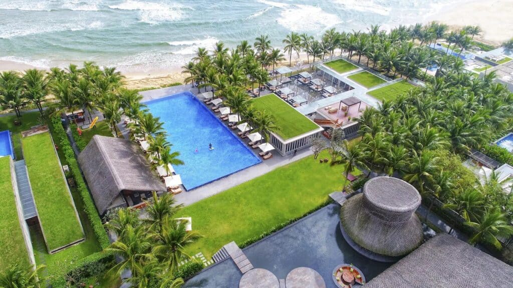 An aerial view of Naman Resort's beachside amenities, featuring a large, vibrant blue pool surrounded by lush palm trees and sun loungers. The pool is adjacent to the sandy beach and the ocean waves, creating a tropical paradise with luxurious and natural elements combined.