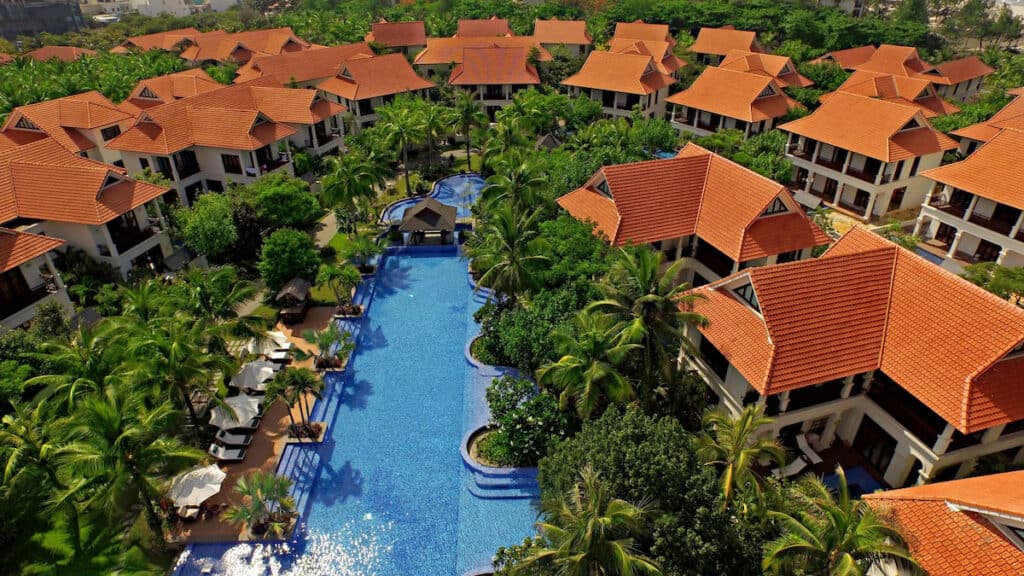 Bird's-eye view of a tropical resort with a snaking blue pool at its center, surrounded by villas with red tile roofs nestled amongst lush greenery