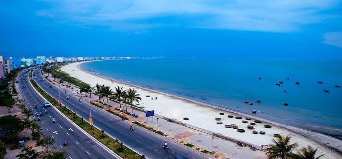 A wide coastal road with palm trees lining one side and a broad sandy beach on the other under a dramatic blue sky. Parked cars dot the beach area, with calm sea waters stretching to the horizon