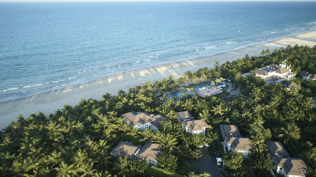 Aerial view of a tranquil beachfront resort with white villas nestled among lush palm trees, adjacent to a serene beach with gentle waves lapping at the shore