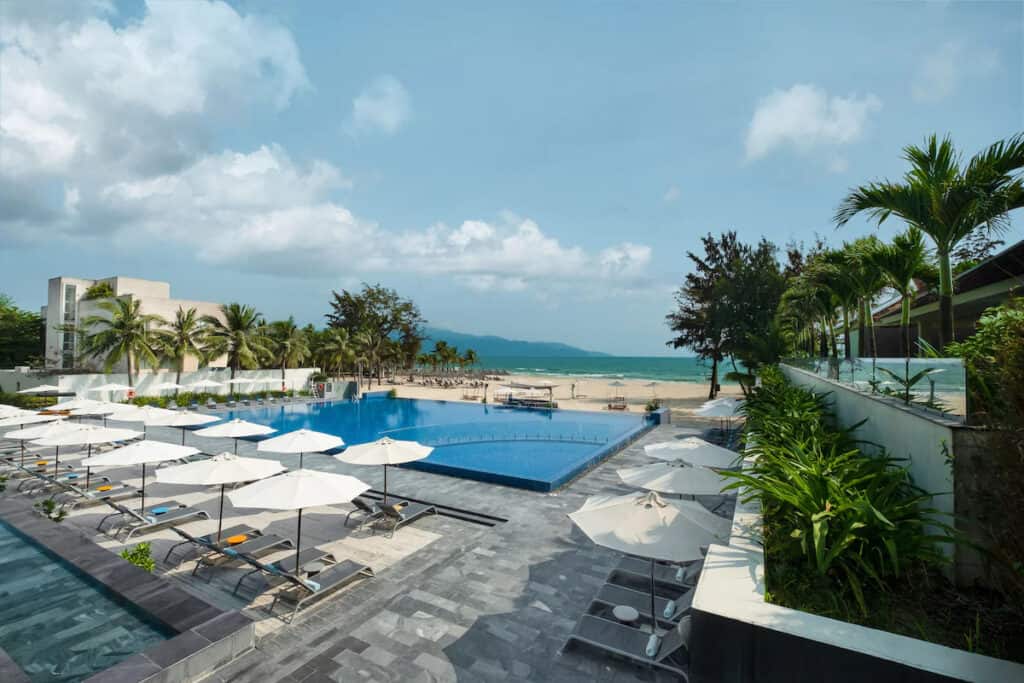 Expansive beachside resort pool area with multiple white umbrellas and sun loungers, offering a refreshing outdoor escape. The pool's edge seamlessly meets the sky, while tropical palms and lush foliage add to the peaceful ambiance, inviting relaxation with a view of the serene beach and distant mountains.