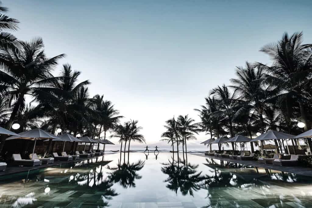 Twilight descends on a serene beachfront infinity pool, with the pool's glassy surface reflecting silhouettes of towering palm trees. White lounge chairs sit under elegant umbrellas, creating a tranquil and luxurious atmosphere, as two people stand at the pool's edge, overlooking the ocean