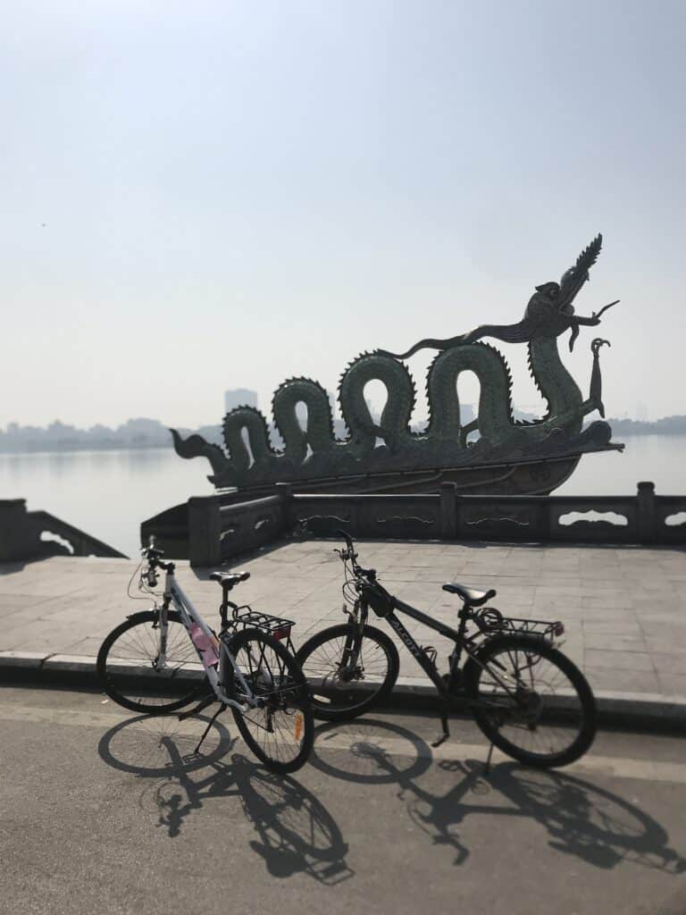 A unique outdoor sculpture of a sea monster with intricate detailing stands prominently by a lakeside, while in the foreground, three parked bicycles cast long shadows on the pavement, suggesting a serene day by the water in Hanoi, Vietnam