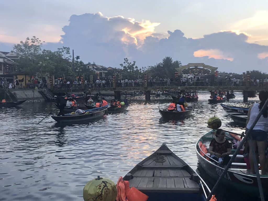 Visitors enjoy a bustling boat ride on a river at dusk with a striking backdrop of a vibrant sunset casting golden hues through dramatic clouds, while onlookers gather along the bridge and riverbank in a quaint, cultural riverside setting in Hoi An, Vietnam