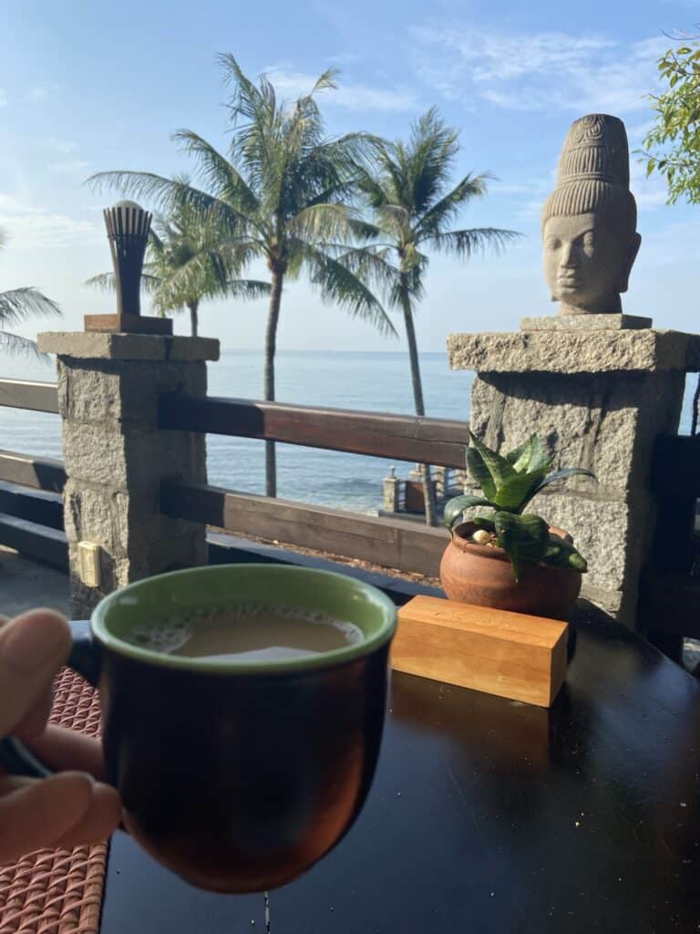 A person's hand holding a dark brown cup with a frothy beverage, in a relaxed outdoor setting featuring a stone sculpture head and lush palm trees against the serene backdrop of a calm sea and clear sky in Phu Quoc, Vietnam.