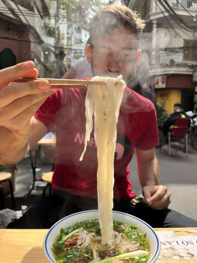 The image shows a man at an outdoor café enjoying a steaming bowl of noodles. He is lifting noodles with chopsticks towards his mouth, with steam visibly rising from the hot soup. The background features an urban street setting, enhancing the casual, lively dining atmosphere.