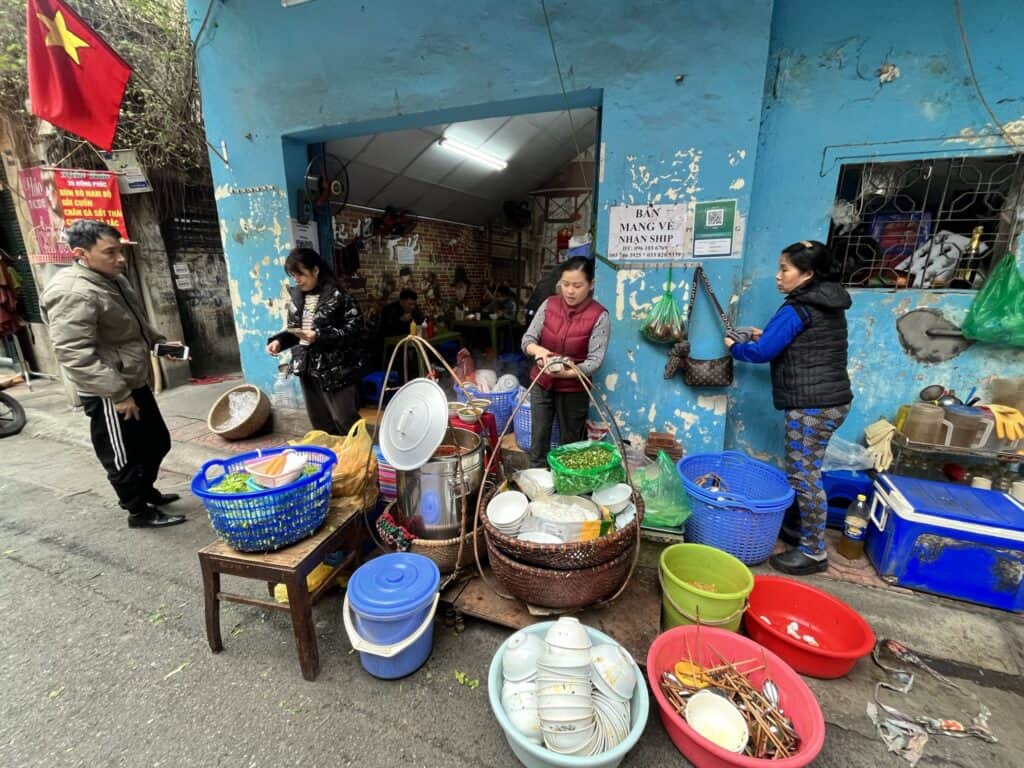 This image captures a vibrant street market scene in a narrow alley with a peeling blue wall. Several women are engaged in different activities: one is selling food items displayed in various colorful baskets and containers on the ground, another is washing dishes in red and pink basins, and a third is cooking. A man in a jacket converses with one of the vendors, adding to the lively atmosphere. In the background, other individuals are dining and socializing in a small eatery, beneath a Vietnamese flag hanging prominently to the left.