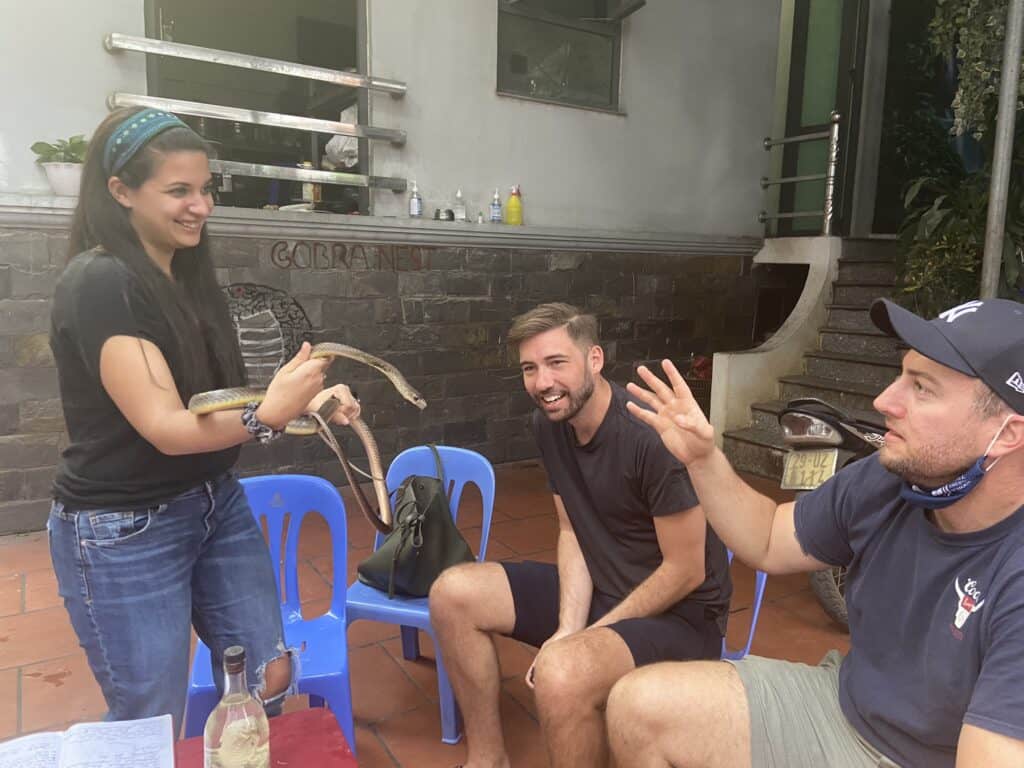 The image shows a casual outdoor gathering where a young woman, smiling, holds a snake in her hands, presenting it to two seated men who appear amused and intrigued. They are in a relaxed, social setting with blue plastic chairs, and behind them, there's a sign that reads "Cobranest," suggesting a wildlife or adventurous theme. The environment has a tropical feel, with visible greenery and a building in the background.