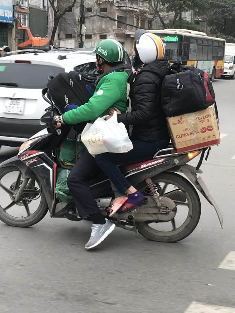 Two people riding a motorbike on a busy street. The driver is wearing a green Grab jacket and helmet, while the passenger behind them is dressed in black with a white and yellow helmet. Both are carrying large backpacks, and the motorbike is loaded with additional bags, including a box of "Biscafun" cookies strapped to the back. The scene is set in an urban environment with other vehicles and buildings in the background.