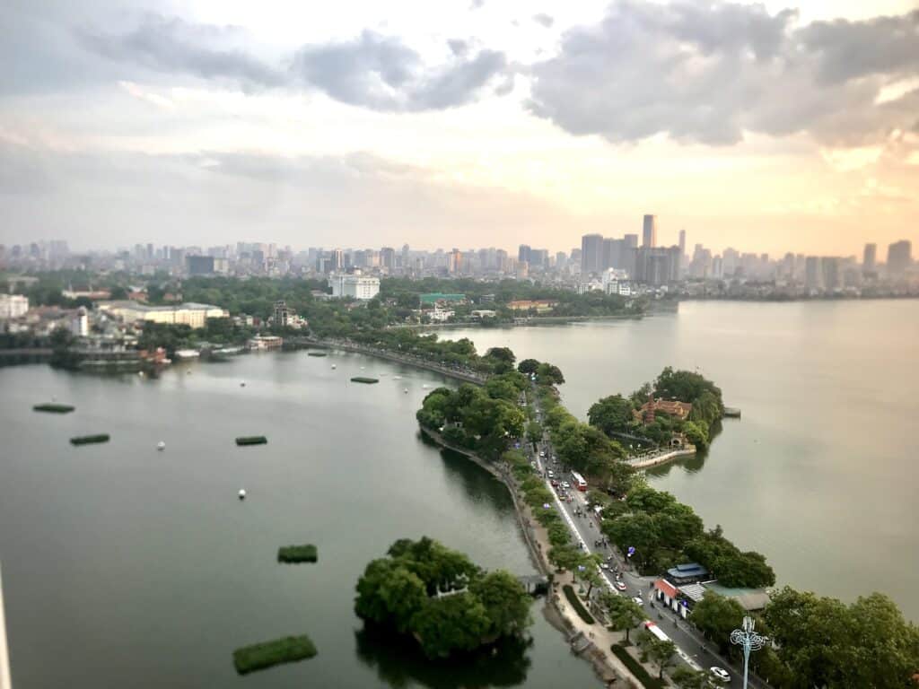 An aerial view of Hanoi's West Lake at sunset, with lush greenery, a winding road, and a small pagoda on an island surrounded by calm water. The city's skyline, featuring modern buildings and a hazy orange sky, stretches across the background.