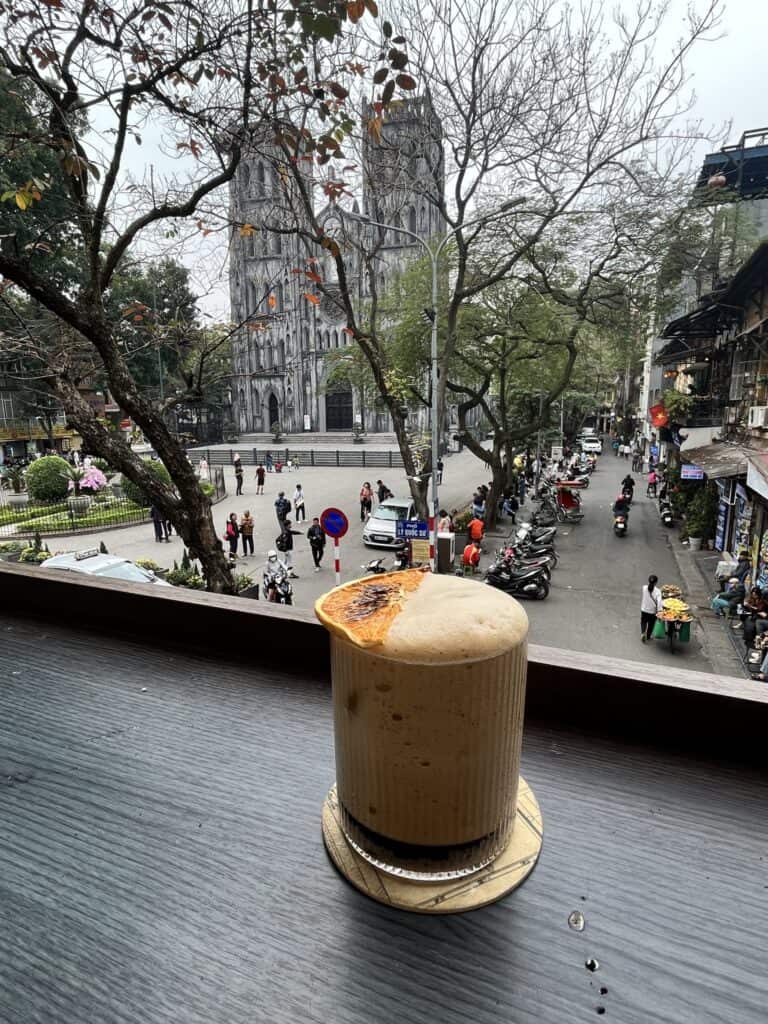 A foamy iced coffee topped with a dried orange slice sits on a table overlooking a bustling Hanoi street with a view of the grand St. Joseph's Cathedral in the background. The scene captures a relaxing café moment, framed by leafless trees and the movement of people and motorbikes below, adding to the vibrant urban atmosphere.