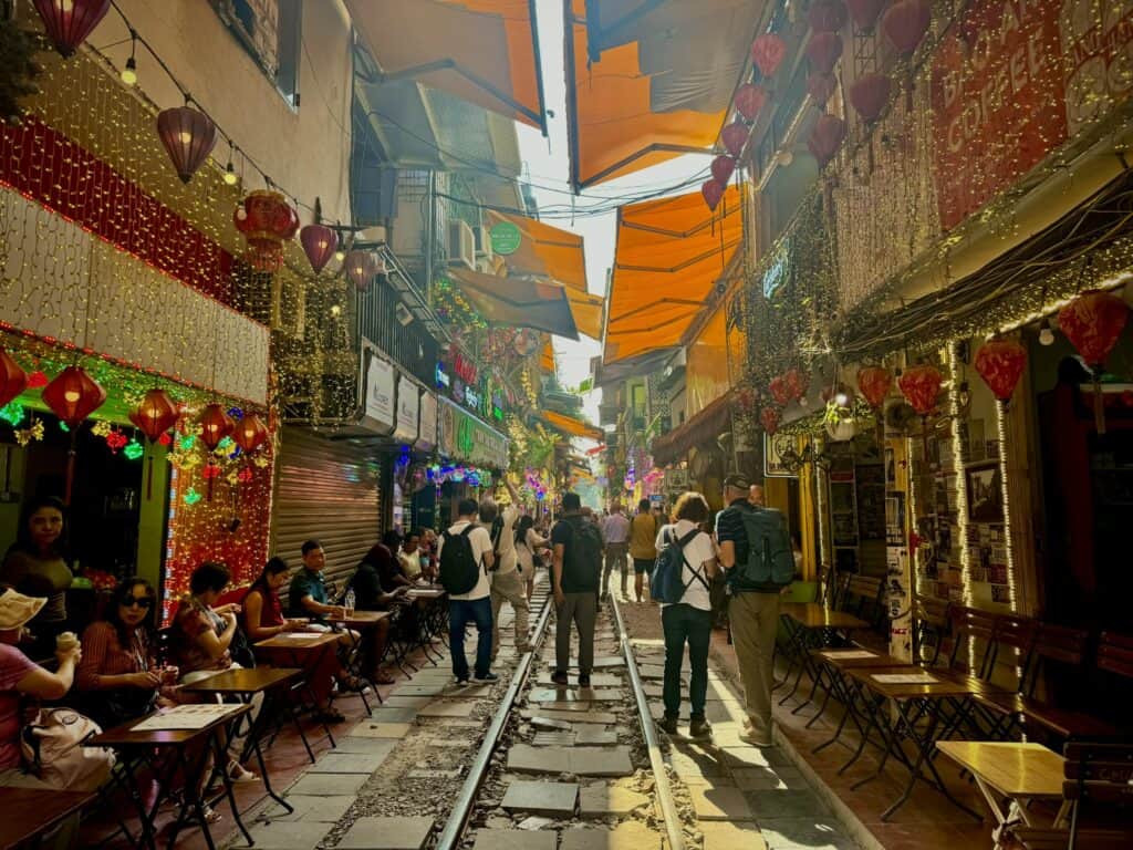 A lively scene on Hanoi's Train Street, with colorful lanterns, string lights, and small cafés lining the narrow railway tracks. Visitors walk along the tracks or sit at outdoor tables, enjoying the vibrant atmosphere under orange awnings on a sunny day.
