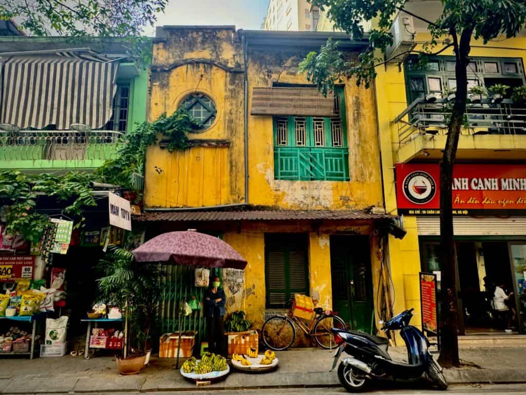 A vibrant street scene in Hanoi, Vietnam, with a rustic yellow building at the center, adorned with green window frames and creeping vines. A street vendor stands under a purple umbrella, selling bananas and other produce, while a motorbike and a bicycle are parked nearby. The scene captures the charm of daily life and the lively mix of colors and textures in the city's architecture.