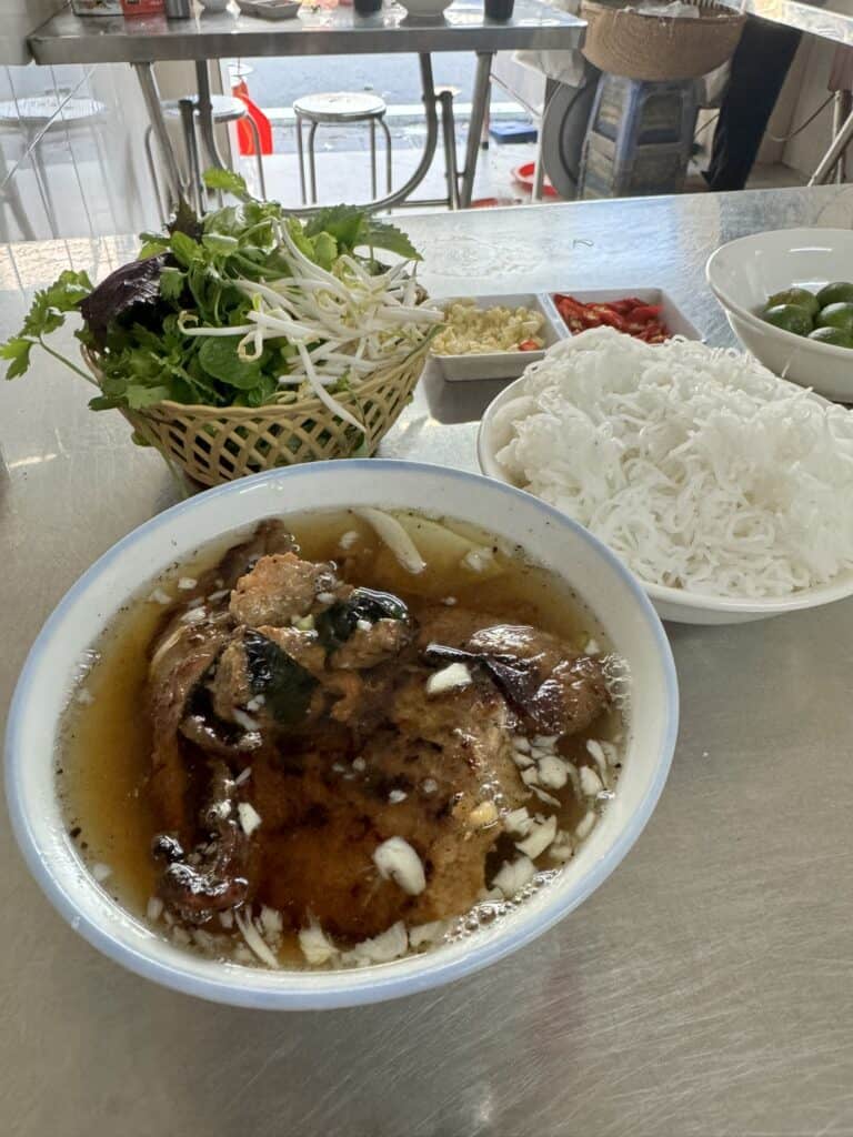 A traditional Vietnamese meal featuring a bowl of bún chả, with grilled pork patties in a light broth, accompanied by a plate of fresh rice noodles, and a basket of mixed herbs and bean sprouts. Side dishes include chopped garlic, red chili slices, and lime, allowing for customizable seasoning. The dish is presented on a metallic table, evoking the casual, authentic setting of a local eatery.