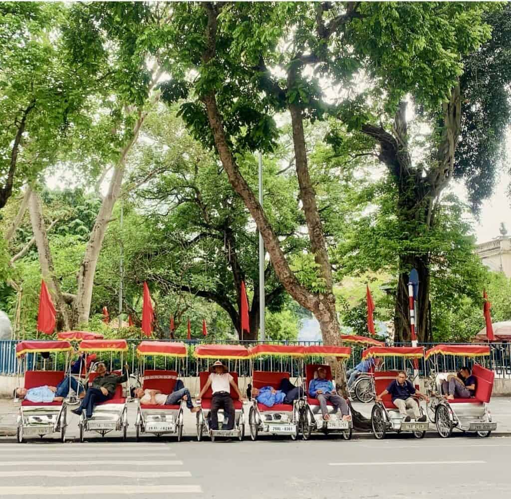 A row of traditional Vietnamese cyclos with red canopies lined up under tall, leafy green trees on a quiet street. Drivers are seen resting or reclining in their seats, with red flags visible in the background, creating a peaceful urban scene.