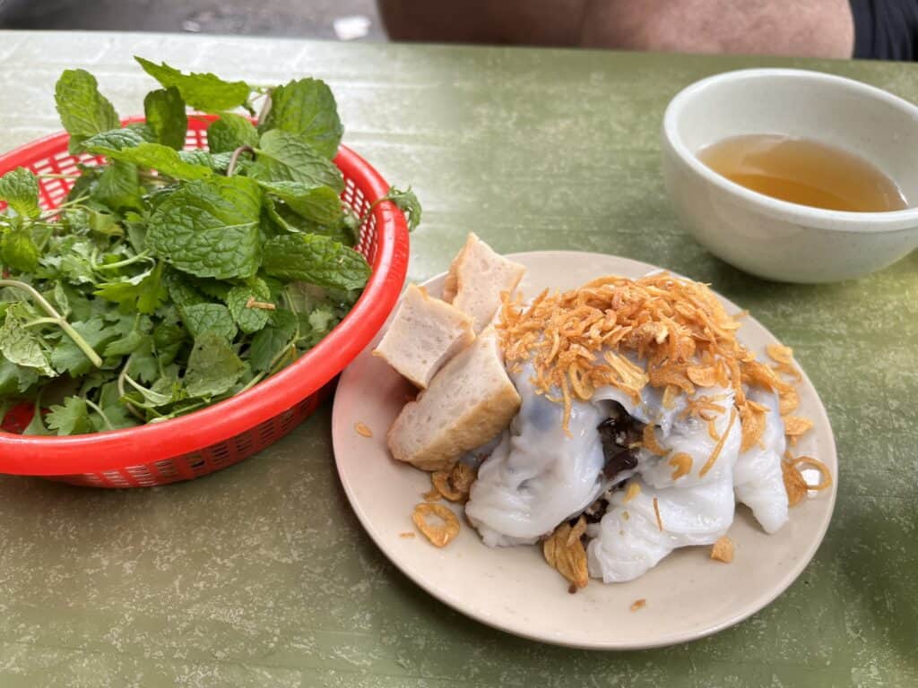 A plate of Vietnamese bánh cuốn (steamed rice rolls) topped with crispy fried shallots and served with slices of Vietnamese sausage on the side. A basket of fresh herbs, including mint and cilantro, and a small bowl of dipping sauce accompany the dish, set on a green table.