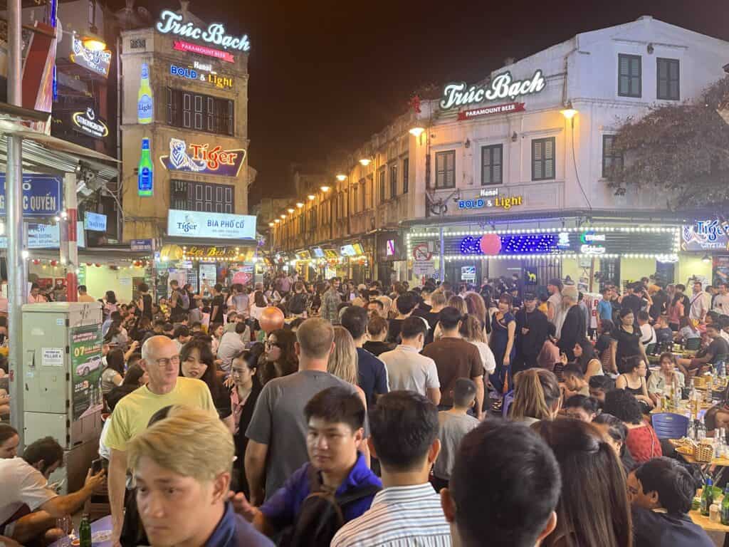 A bustling nightlife scene in Hanoi's Old Quarter, with crowds of people filling the street surrounded by brightly lit signs for beer and restaurants, including 'Trúc Bạch' and 'Tiger Beer.' The lively atmosphere features people dining, drinking, and socializing under the glow of neon lights.