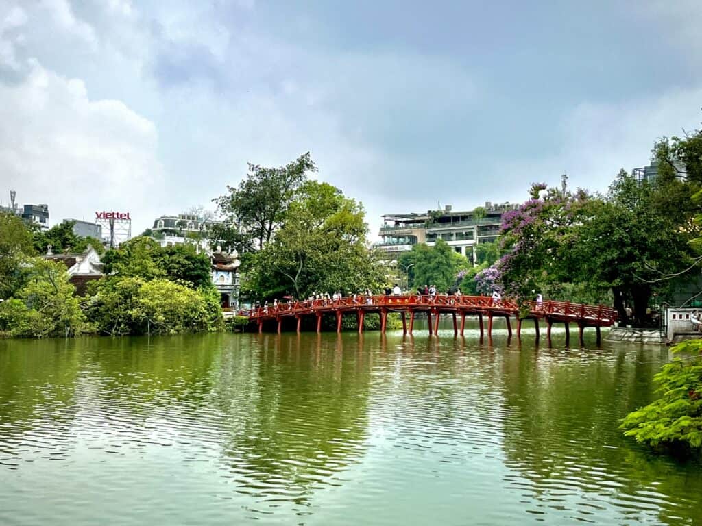 The iconic red wooden bridge, The Huc Bridge, spans a calm green lake at Hoan Kiem Lake in Hanoi, Vietnam. Surrounded by lush trees, blooming purple flowers, and historic buildings, the scene exudes tranquility under a partly cloudy sky.