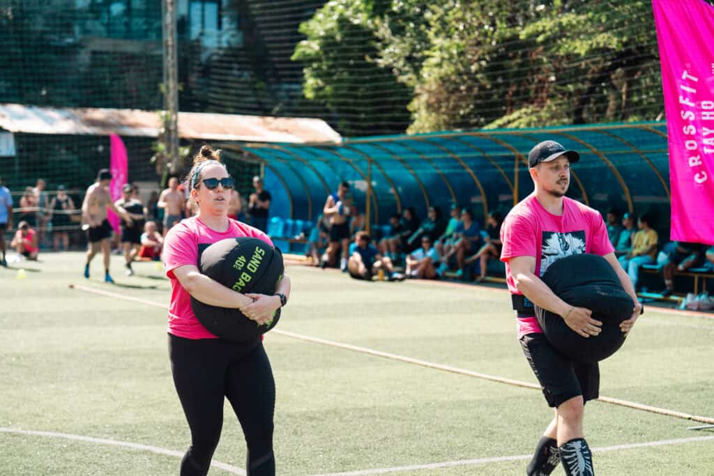 Two participants in a CrossFit competition carrying heavy sandbags across a field, both wearing bright pink shirts. Spectators sit in the background under a shaded seating area, with vibrant pink banners displaying 'CrossFit Tay Ho' adding energy to the outdoor event.