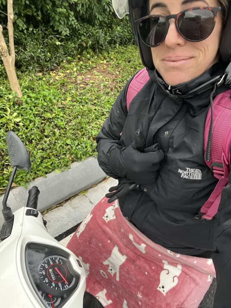 A person wearing a helmet, sunglasses, and a black North Face jacket sits on a scooter, wrapped in a red blanket with white cat patterns for warmth. The background features a green hedge and a sidewalk, indicating a chilly outdoor setting.