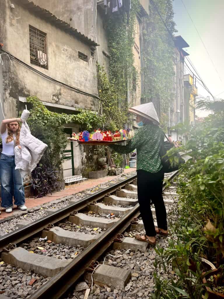 A street vendor wearing a conical hat and green patterned shirt stands on railway tracks, holding a tray of colorful goods. The setting features a residential building covered in greenery, while another person is seen in the background posing for a photo near the tracks.