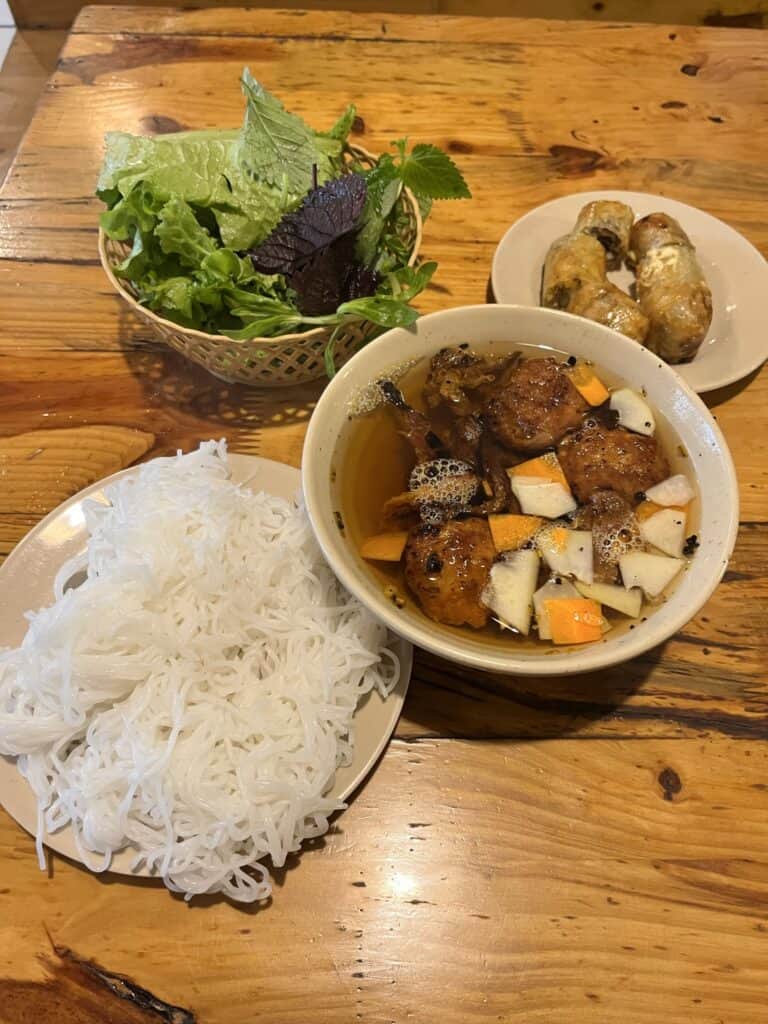 A traditional Vietnamese meal featuring a bowl of bún chả, served with grilled pork patties in a tangy-sweet broth with pickled carrots and daikon. To the left, a plate of fresh, white vermicelli noodles is neatly arranged. A small basket of fresh herbs, including lettuce and perilla leaves, sits in the background, while a separate plate holds two golden-brown fried spring rolls. The rustic wooden table enhances the warm, inviting presentation of the dish.