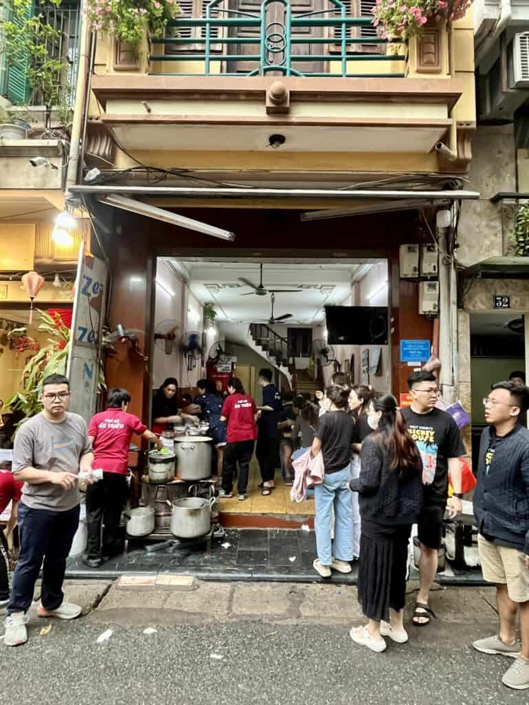 Exterior view of a busy Vietnamese street food restaurant with an open entrance. Customers line up outside while staff in red uniforms prepare food at large metal pots near the doorway. The building features a balcony with potted flowers, blending a residential and commercial vibe