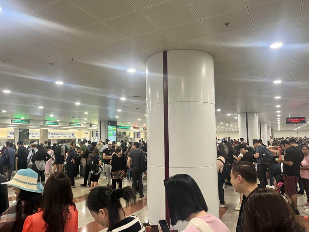 Crowded airport terminal with people gathered near counters under a brightly lit ceiling. Many individuals are standing in lines or waiting, some holding documents, while others appear to be checking their phones. Bank counters and service signs are visible in the background