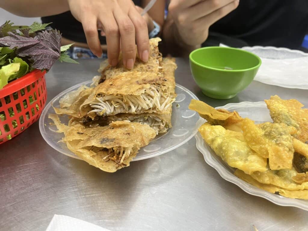 A plate of crispy bánh xèo, a Vietnamese savory rice flour pancake, folded and filled with bean sprouts, mushrooms, and possibly meat, with a diner using their hands to pick up a piece. To the right, a plate of golden fried wontons sits beside a small green dipping bowl. A basket of fresh herbs, including perilla leaves and lettuce, is on the table, along with rice paper for wrapping. The setting appears to be a casual dining spot with a stainless steel table.