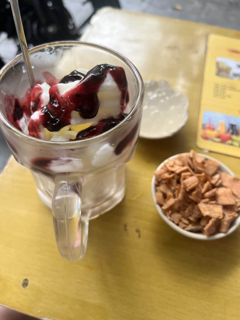 A glass mug filled with creamy yogurt, drizzled with a dark berry sauce, possibly blackcurrant or blueberry, with a spoon resting inside. Beside it, a small bowl of crispy, golden-brown coconut flakes sits on a yellow table. In the background, there’s another dish with a translucent jelly dessert and a laminated menu featuring colorful drink options. The setting suggests an outdoor or casual café atmosphere.