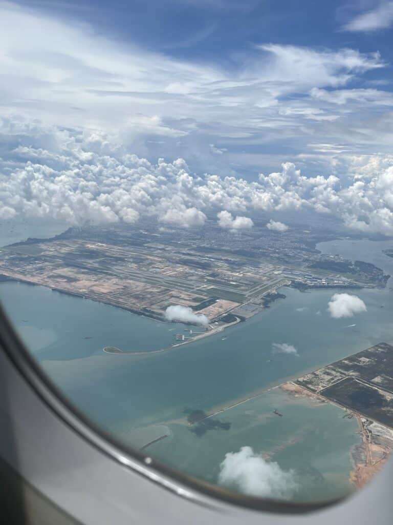 Aerial view from an airplane window showing a coastline with a blend of natural landscape and developed areas. The sky is filled with fluffy clouds under a bright blue sky, while the water below reflects shades of blue and green. Runways, small islands, and coastal infrastructure are visible along the shoreline.