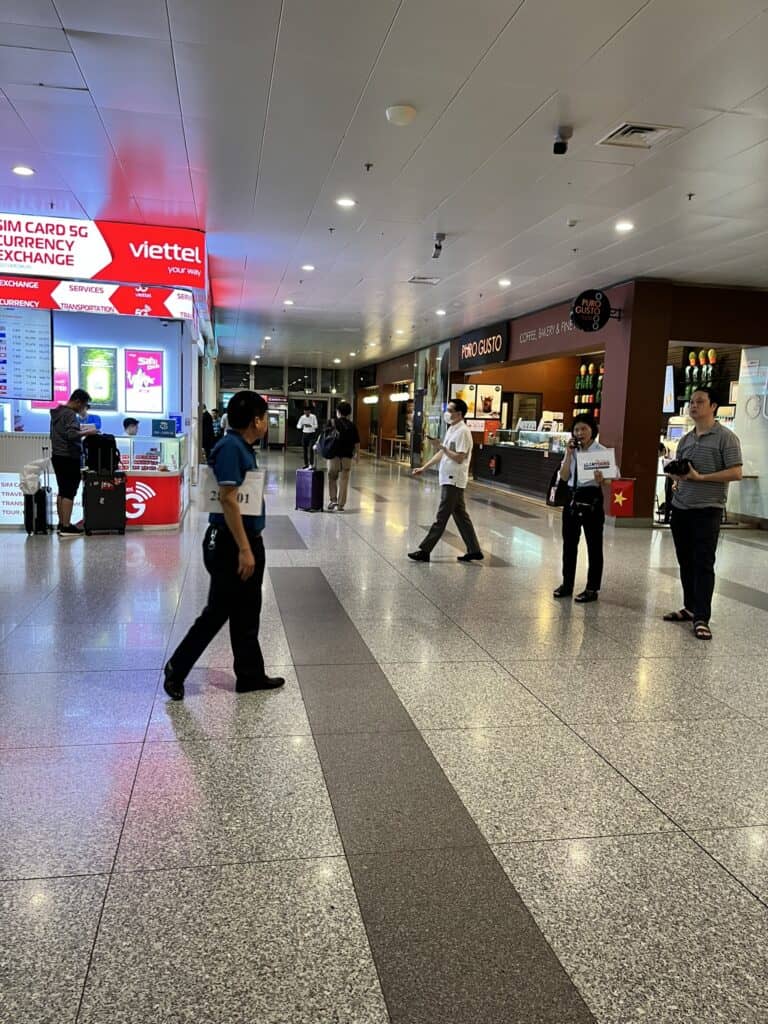 Airport terminal scene with a SIM card and currency exchange booth on the left displaying a red "Viettel" sign. Several travelers and staff are walking or standing, some with luggage. On the right, a café called "PHO GUSTO" offers coffee and bakery items. The floor is polished with reflective tiles under bright overhead lighting